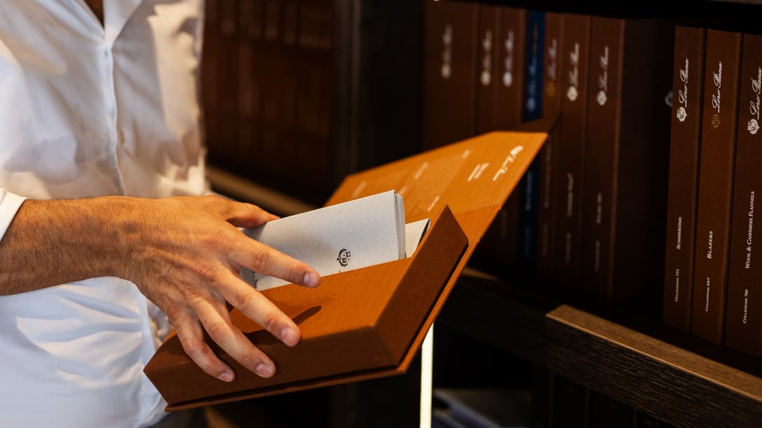 Eine Hand zieht Stoffmuster aus einer aufgeklappten Loro-Piana-Box, dahinter ein Regal mit Loro-Piana-Stoffbüchern