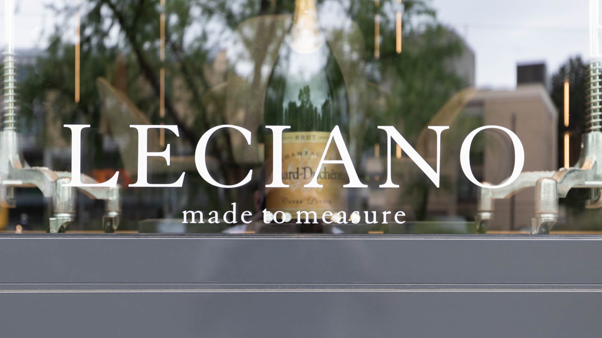 Schriftzug LECIANO – made to measure auf der Schaufensterscheibe, dahinter eine Flasche Champagner